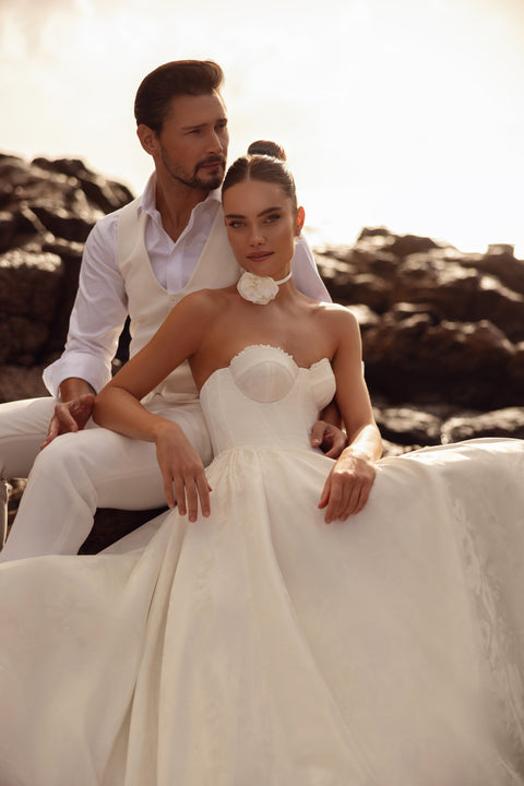 Suknia ślubna Cholly w klasycznym stylu z gorsetem i rozkloszowanym dołem, prezentowana w romantycznej sesji na tle morskich skał.